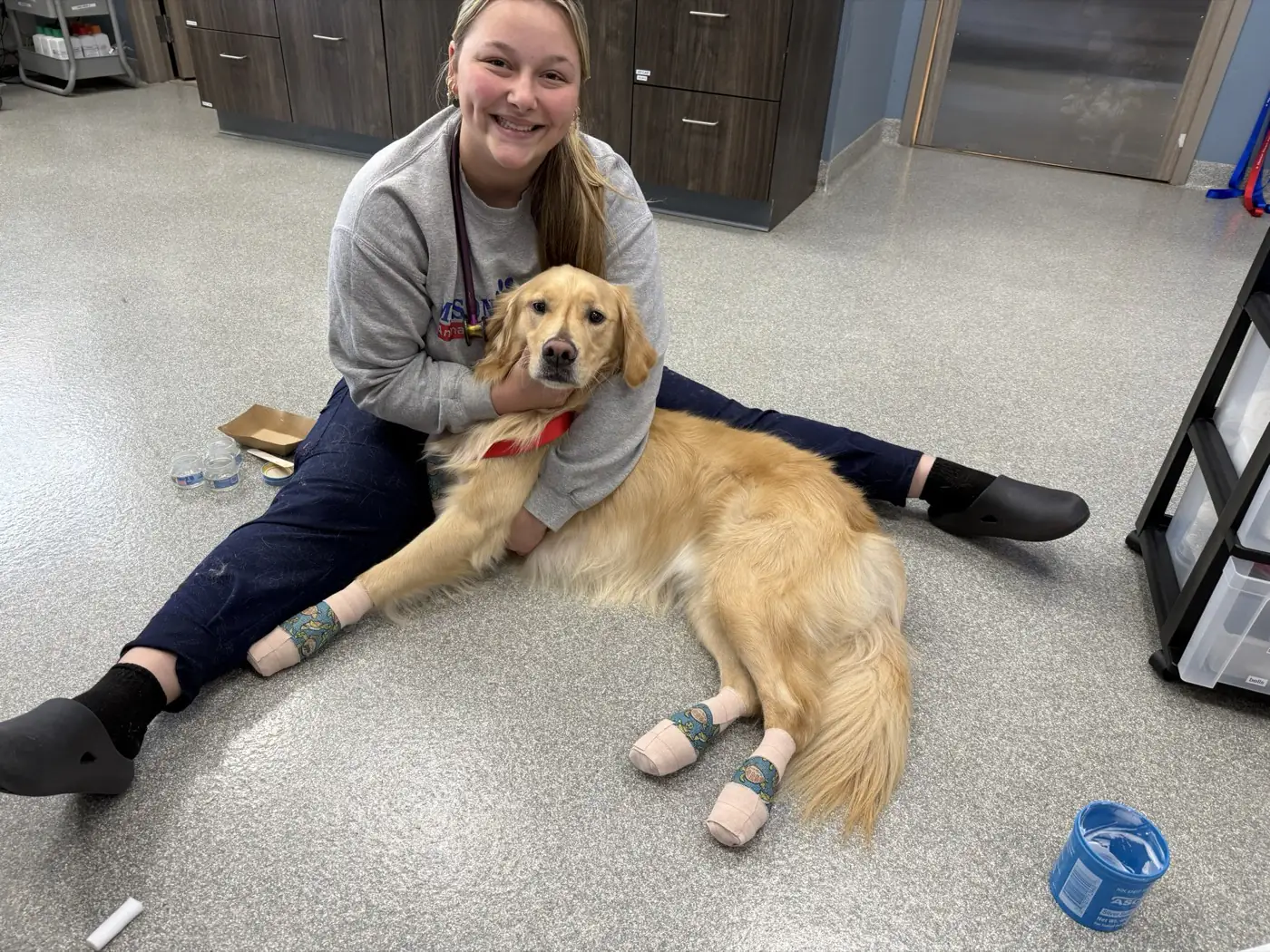 A smiling team member sitting on the treatment room floor hugging a golden retriever with bandaged paws.